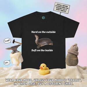 May include: A black t-shirt featuring a turtle graphic with the text Hard on the outside and Soft on the inside. Above the shirt is a sign reading Official uniform of people who look tough but cry at movie trailers. Includes a hammer and anvil.