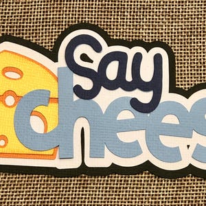 May include: Say Cheese! A blue and white paper craft with the words "Say Cheese" and a yellow cheese wedge.