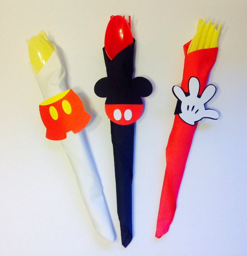 Op de afbeelding: Drie decoratieve servethouders met Mickey Mouse-thema's. E&eacute;n heeft geel bestek en een rode broek, een andere een rode lepel en een Mickey Mouse-hoofd, en de laatste heeft gele vorken en een witte handschoen.