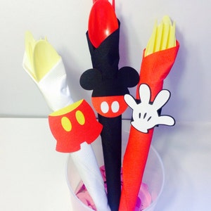 Op de afbeelding: Een doorzichtige plastic beker gevuld met feestartikelen met Mickey Mouse-thema. De benodigdheden omvatten een rode lepel en vork, een gele vork en een rode servet met een witte handschoen uitsparing. De benodigdheden zijn versierd met Mickey Mouse-thema uitsparingen.