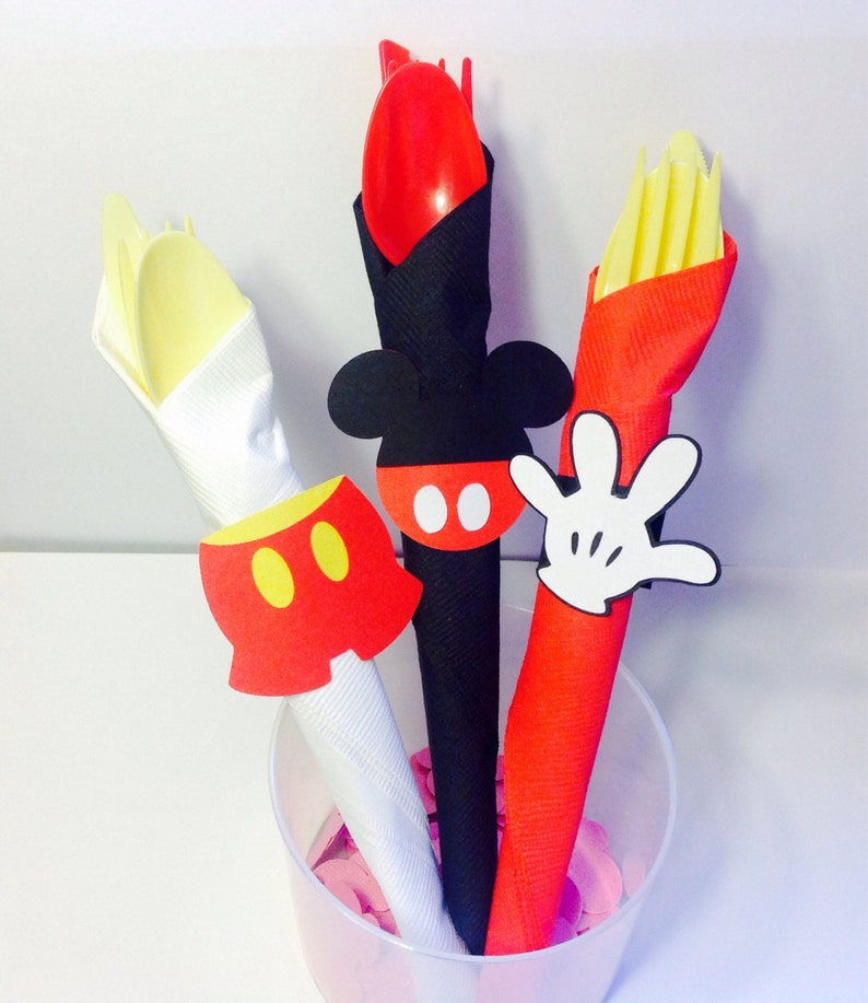 Op de afbeelding: Een doorzichtige plastic beker gevuld met feestartikelen met Mickey Mouse-thema. De benodigdheden omvatten een rode lepel en vork, een gele vork en een rode servet met een witte handschoen uitsparing. De benodigdheden zijn versierd met Mickey Mouse-thema uitsparingen.