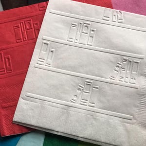 May include: A white paper napkin with an embossed design of bookshelves with books on them. The napkin is layered on top of other napkins in various colors, including red, blue, green, and pink.