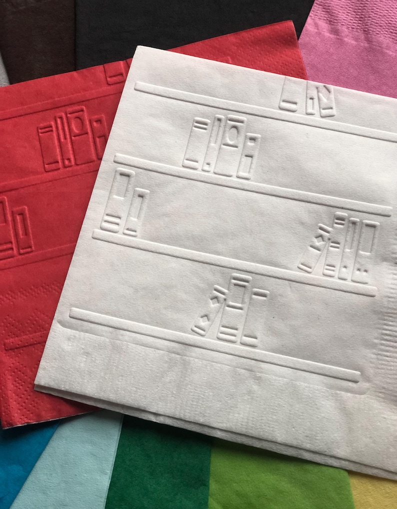 Puede incluir: Una servilleta de papel blanca con un dise&ntilde;o en relieve de estanter&iacute;as con libros. La servilleta est&aacute; superpuesta sobre otras servilletas de varios colores, incluyendo rojo, azul, verde y rosa.