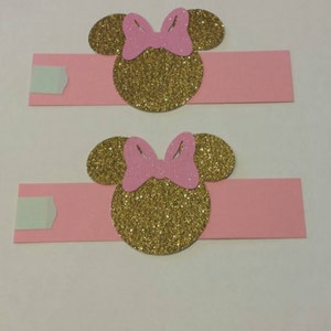 K&ouml;nnte beinhalten: Zwei rosa Serviettenringe mit goldenen Glitzer-Minnie-Maus-K&ouml;pfen. Jeder Ring hat eine rosa Schleife auf dem Kopf.