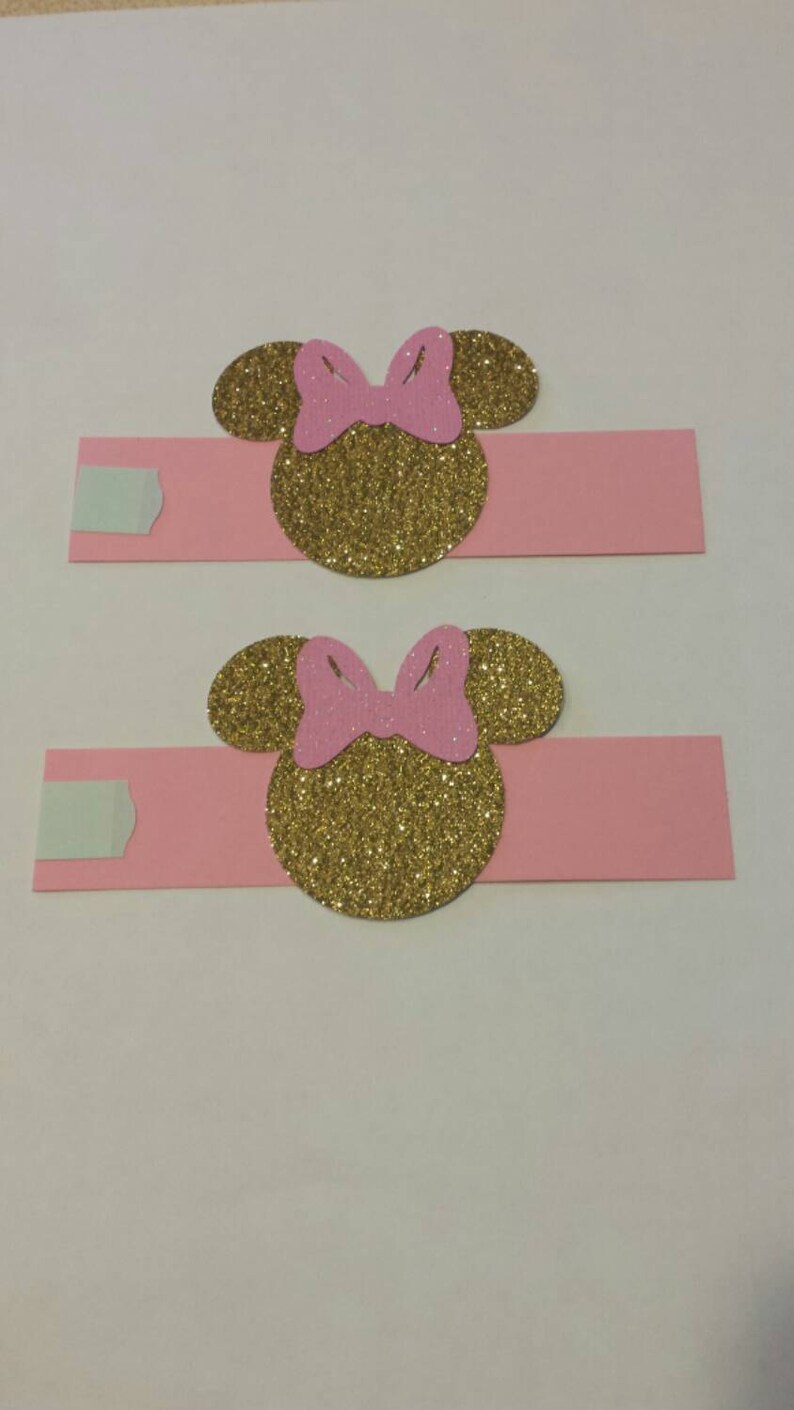 K&ouml;nnte beinhalten: Zwei rosa Serviettenringe mit goldenen Glitzer-Minnie-Maus-K&ouml;pfen. Jeder Ring hat eine rosa Schleife auf dem Kopf.