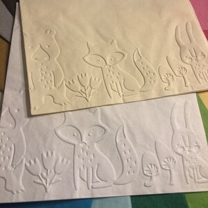 Puede incluir: Dos servilletas de papel blancas con dise&ntilde;os en relieve de un zorro, un oso y un conejo. La servilleta de arriba es de color crema y la servilleta de abajo es blanca.