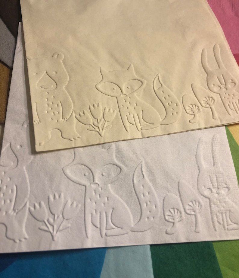 Puede incluir: Dos servilletas de papel blancas con dise&ntilde;os en relieve de un zorro, un oso y un conejo. La servilleta de arriba es de color crema y la servilleta de abajo es blanca.