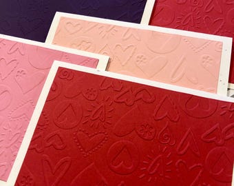 May include: A collection of embossed paper cards in various shades of pink, red, and purple. Each card features a repeating pattern of hearts and floral designs, with a white border. The cards are arranged on a patterned background.