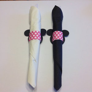 Op de afbeelding: Twee opgerolde servetten, &eacute;&eacute;n witte en &eacute;&eacute;n zwarte, vastgehouden door decoratieve servetringen. Elke ring heeft een roze stippenband en zwarte muizenoren. De servetten liggen op een wit oppervlak.