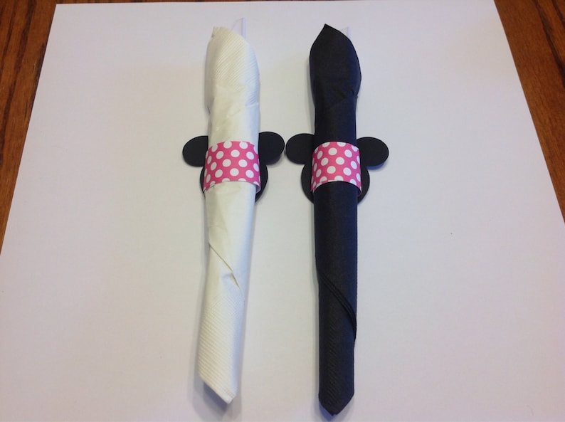 Op de afbeelding: Twee opgerolde servetten, &eacute;&eacute;n witte en &eacute;&eacute;n zwarte, vastgehouden door decoratieve servetringen. Elke ring heeft een roze stippenband en zwarte muizenoren. De servetten liggen op een wit oppervlak.