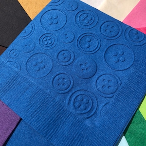 May include: A blue paper napkin with a repeating pattern of embossed buttons. The napkin is surrounded by other colored paper napkins.