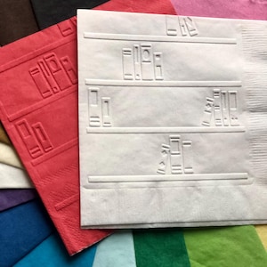 Puede incluir: Una servilleta de papel blanca con un dise&ntilde;o de estanter&iacute;a. La servilleta tiene un patr&oacute;n en relieve de libros en estantes. La servilleta est&aacute; rodeada de otras servilletas de varios colores.