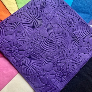 May include: Purple paper napkin with an embossed design of seashells, starfish, and swirls. The napkin is on top of a stack of colorful paper napkins.