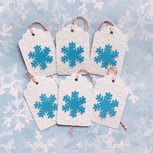 May include: Set of six white gift tags with blue snowflake embellishments. Each tag has a hole punched at the top and is tied with red and white twine.