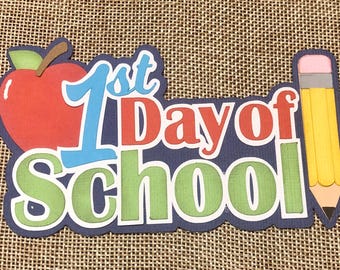 May include: A colorful paper craft with the words "1st Day of School" in red, blue, and green letters. A red apple and a yellow pencil are on either side of the words.