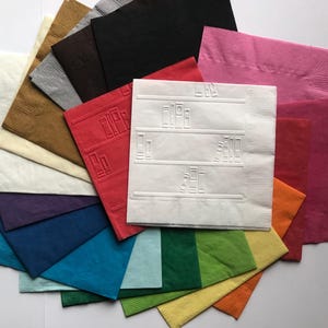 Puede incluir: Una pila de 15 servilletas de papel en varios colores, incluyendo rojo, azul, verde, amarillo y blanco. Las servilletas est&aacute;n dobladas por la mitad y tienen una superficie texturizada. Una servilleta tiene un dise&ntilde;o blanco en relieve con l&iacute;neas y cuadrados.
