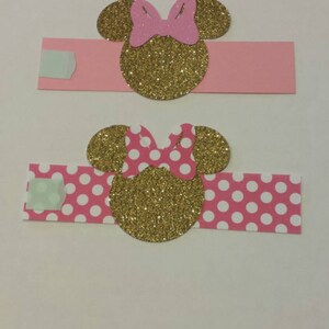 K&ouml;nnte beinhalten: Zwei rosa und goldene Minnie Mouse Serviettenringe. Ein Ring hat einen einfarbigen rosa Hintergrund und der andere einen rosa Hintergrund mit wei&szlig;en Punkten.