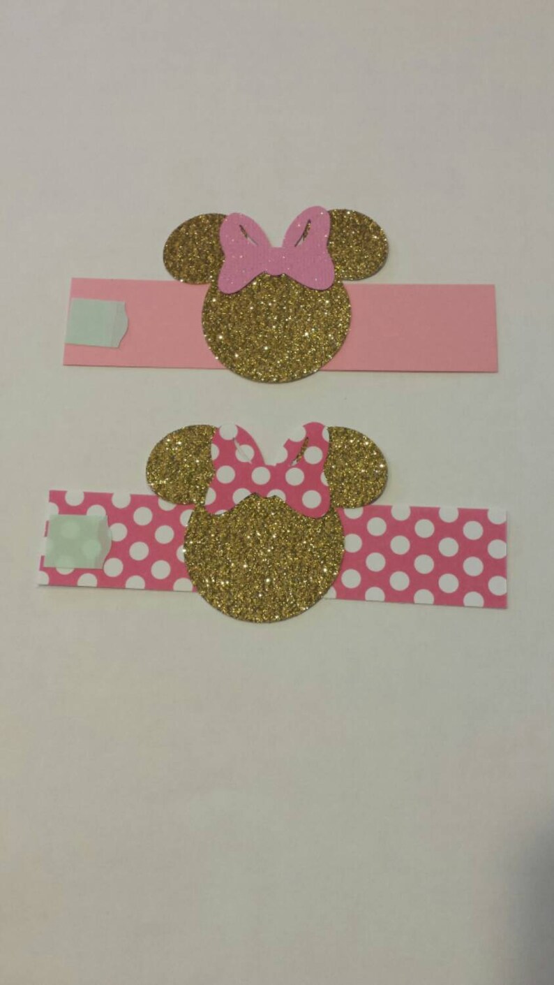 K&ouml;nnte beinhalten: Zwei rosa und goldene Minnie Mouse Serviettenringe. Ein Ring hat einen einfarbigen rosa Hintergrund und der andere einen rosa Hintergrund mit wei&szlig;en Punkten.