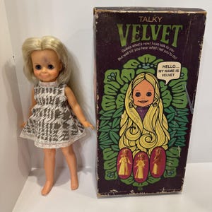 May include: A vintage "Talky Velvet" doll with blonde hair and a patterned dress stands beside its original box. The box features a cartoon illustration of the doll and the text "HELLO... MY NAME IS VELVET". The doll is a retro toy.
