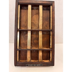 Puede incluir: Una bandeja de tipógrafo de madera vintage con compartimentos y divisores de latón. Los números 2, 3, 6, 12, 18, 24, 30 y 36 son visibles. El texto "SPACES AND QUADS" está impreso en una pequeña placa de metal en la parte inferior. La bandeja es de color marrón oscuro.