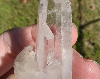 May include: A clear quartz crystal with a pointed tip and a rough, textured surface. The crystal is held in a hand against a blurred green background. The crystal's facets catch the light, showcasing its transparency.