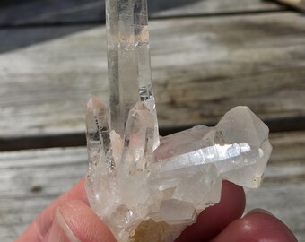 May include: A cluster of clear quartz crystals, with a large central point and several smaller points branching out. The crystals are translucent, with visible internal structures. The quartz is held in a hand.