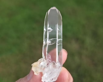 May include: A clear quartz crystal with a pointed tip. The crystal is held in a hand against a blurred green background. The base of the crystal has a rough, white and yellow appearance.