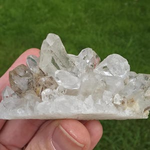 May include: A cluster of clear quartz crystals with a rough, natural texture. The crystals vary in size and shape, with some appearing more transparent than others. The base of the cluster is a light, off-white color, and the background is a blurred green.