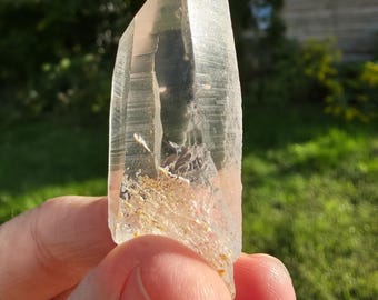 May include: A clear quartz crystal point, held in a hand, with a natural, rough base and a polished, faceted tip. The crystal has internal inclusions and a slightly pinkish hue, with visible lines and textures. The background is blurred green foliage.