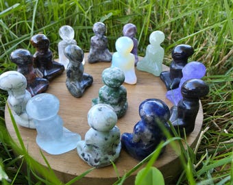 May include: A collection of small, seated stone figurines in various colors, including blue, green, and purple, arranged on a wooden surface. The figurines are set against a backdrop of green grass.