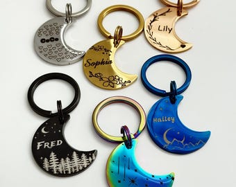 May include: A collection of crescent moon-shaped keychains in various colors, including silver, gold, black, blue, and iridescent. Each keychain features a different design, such as hearts, floral patterns, and names like "Fred" and "Rosie".
