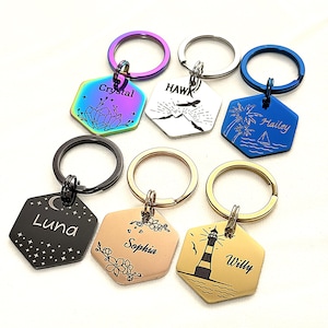 May include: A collection of hexagonal keychains with various designs and names. Keychains feature a variety of colors, including iridescent, black, blue, and gold. Designs include a crystal, a hawk, palm trees, a moon and stars, flowers, and a lighthouse. Each keychain has a metal ring.
