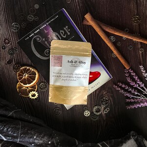 May include: A package labeled "Ash & Alloy" rests on a book titled "Cinder" with a dark wooden background. Dried orange slices, cinnamon sticks, and decorative gears surround the package and book. The package has a quote on it.