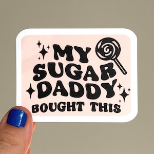 May include: A rectangular sticker with a light pink background and a white border. The sticker features black text that reads MY SUGAR DADDY BOUGHT THIS in a bold, wavy font, accompanied by a black lollipop graphic and several small star icons.