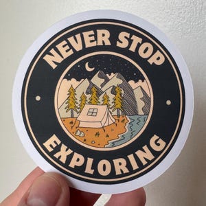 Può includere: Adesivo rotondo con bordo nero e la scritta "NEVER STOP EXPLORING" in crema. Il centro presenta una scena di campeggio con montagne, alberi, una tenda e una luna crescente su uno sfondo stellato.