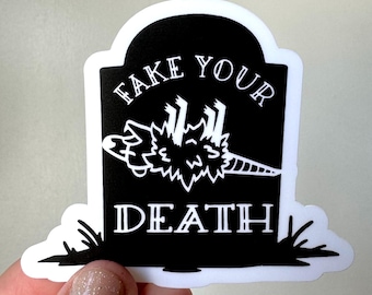 May include: A black and white sticker in the shape of a tombstone. The text "FAKE YOUR DEATH" is at the top and "DEATH" at the bottom. A cartoonish, upside-down creature is in the center.