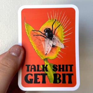 May include: A rectangular sticker with a red background featuring a Venus flytrap with a fly inside. The flytrap is yellow and green. The sticker has the text "TALK SHIT GET BIT" in black letters.