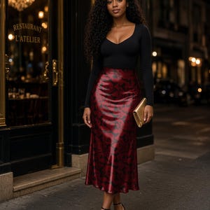 May include: A long-sleeved black bodysuit with a scoop neckline paired with a high-waisted, calf-length satin skirt in deep red featuring a black abstract pattern. The outfit includes black strappy high-heeled sandals and a small gold clutch.