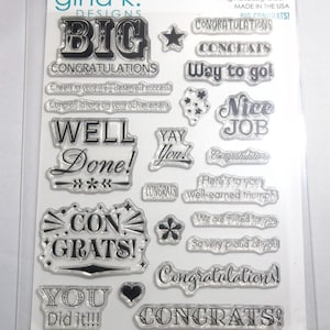 May include: Clear photopolymer stamps with congratulatory phrases like "Big Congratulations," "Well Done," and "Nice Job." The stamps include stars, hearts, and floral designs. The package is labeled "gina k. DESIGNS."