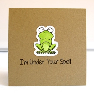 May include: A square, brown card with a green frog sticker and the text "I'm Under Your Spell" in black. The frog is light green with black and white details.