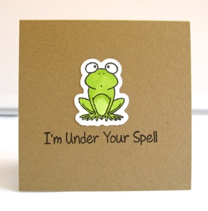May include: A square, brown card with a green frog sticker and the text "I'm Under Your Spell" in black. The frog is light green with black and white details.