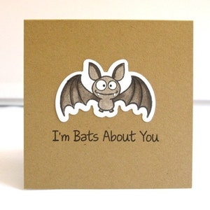 May include: A square, kraft paper greeting card featuring a cartoon bat illustration. The bat is gray with large eyes and a smiling expression. The text "I'm Bats About You" is printed below the bat.