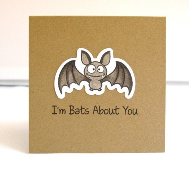 May include: A square, kraft paper greeting card featuring a cartoon bat illustration. The bat is gray with large eyes and a smiling expression. The text "I'm Bats About You" is printed below the bat.