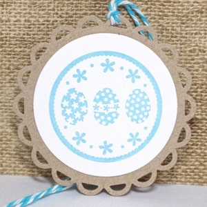 May include: A round, brown paper gift tag with a white circle in the center. The circle has a blue border with white polka dots and three blue eggs with white polka dots and flower patterns. The tag has a scalloped edge and is tied with a blue and white twine.