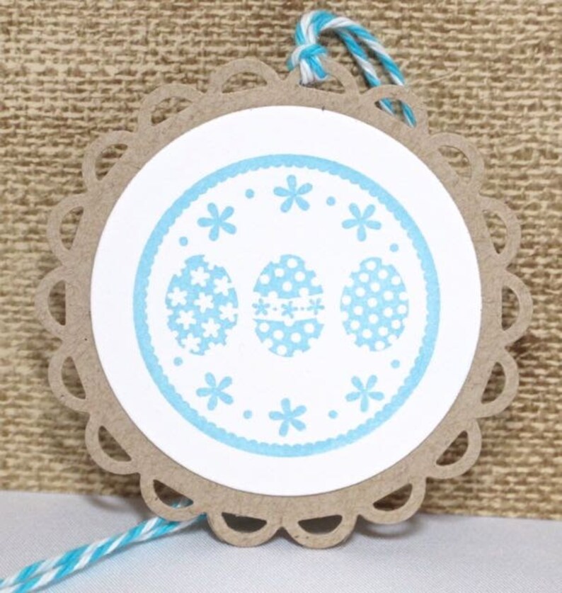 May include: A round, brown paper gift tag with a white circle in the center. The circle has a blue border with white polka dots and three blue eggs with white polka dots and flower patterns. The tag has a scalloped edge and is tied with a blue and white twine.