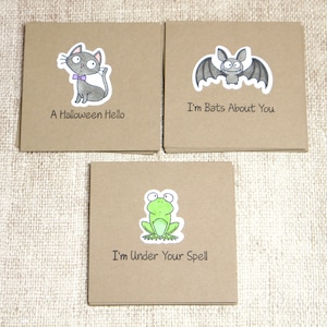 May include: Three square greeting cards with cartoon illustrations. One card features a gray cat with a purple bow tie and the text "A Halloween Hello." Another card has a bat with the text "I'm Bats About You." The third card shows a frog with the text "I'm Under Your Spell."