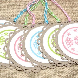 May include: Six round, brown paper gift tags with scalloped edges. Each tag has a white circle with a different pastel colored design of eggs and flowers. The tags are tied with twine.