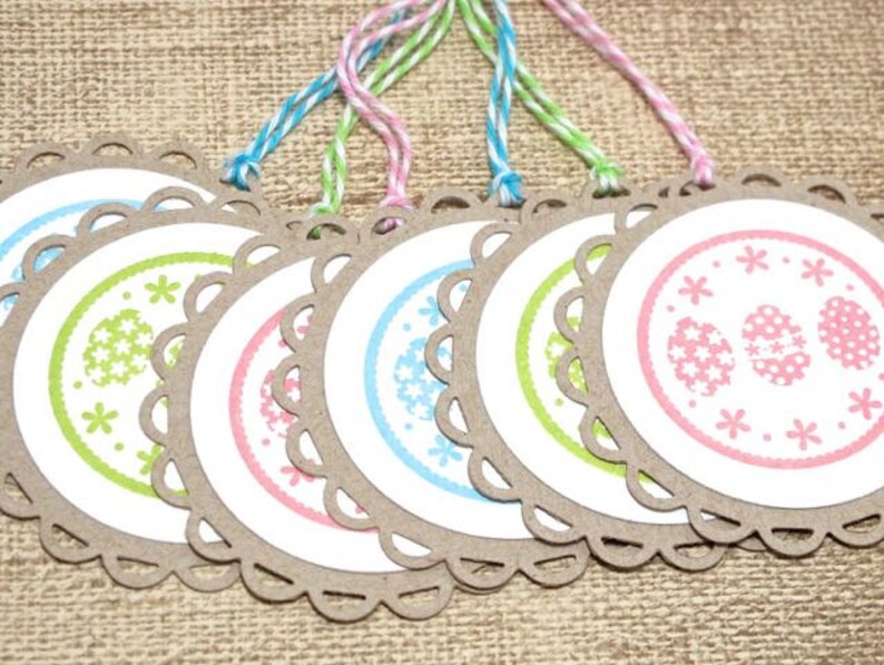 May include: Six round, brown paper gift tags with scalloped edges. Each tag has a white circle with a different pastel colored design of eggs and flowers. The tags are tied with twine.