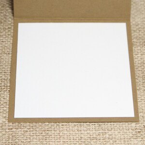 May include: A blank greeting card with a white interior and a brown, textured exterior. The card is open, revealing the blank white space for a personalized message. The card is set against a neutral background.