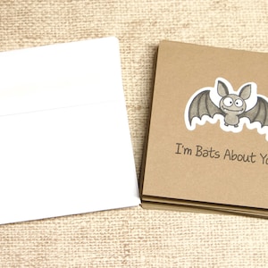 May include: A stack of brown cards with a cartoon bat illustration and the text "I'm Bats About You" next to a stack of white envelopes. The bat is gray with a white outline. The cards and envelopes are on a textured, tan surface.